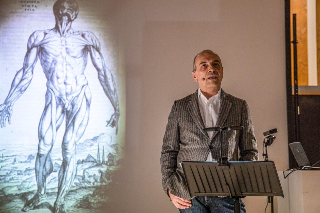 Il fisico Diego Bettoni durante lo spettacolo La fisica immagina il corpo. Alle sue spalle proiettato un disegno anatomico che mostra i muscoli di una figura umana.