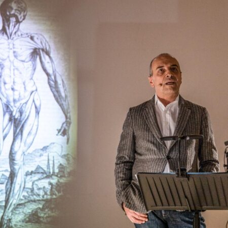 Il fisico Diego Bettoni durante lo spettacolo La fisica immagina il corpo. Alle sue spalle proiettato un disegno anatomico che mostra i muscoli di una figura umana.
