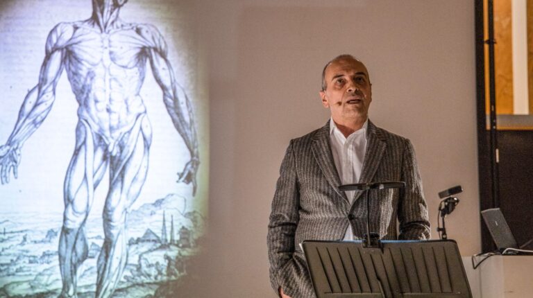 Il fisico Diego Bettoni durante lo spettacolo La fisica immagina il corpo. Alle sue spalle proiettato un disegno anatomico che mostra i muscoli di una figura umana.